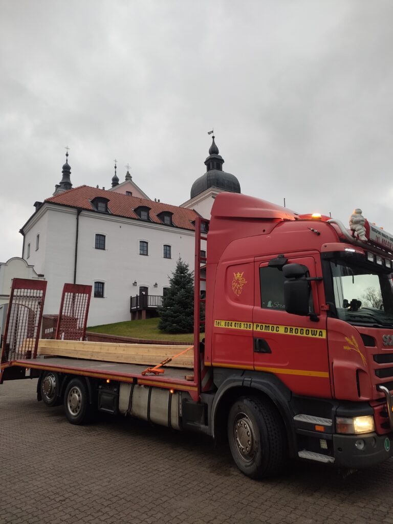 Czerwona laweta firmy Walenda Usługi Sprzętowe na tle zabytkowego budynku w Suwałkach, oferująca wynajem maszyn i usługi transportowe.